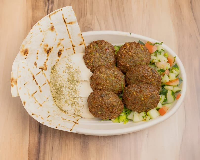 A white bowl filled with falafel, hummus, pita bread, and a cucumber and tomato salad.