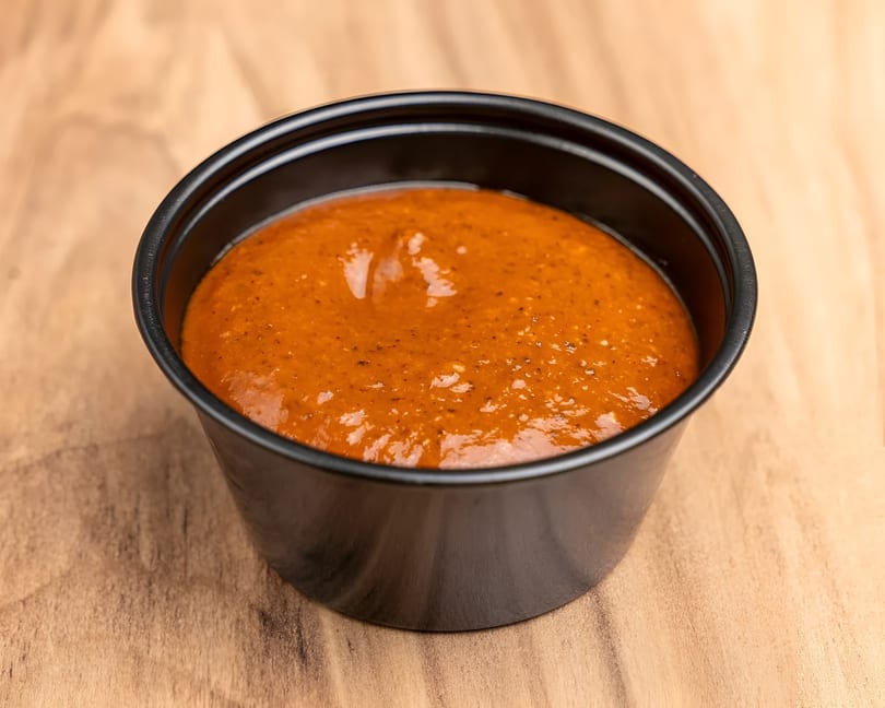 A black plastic cup filled with a vibrant orange-red sauce, speckled with spices, on a wooden surface.