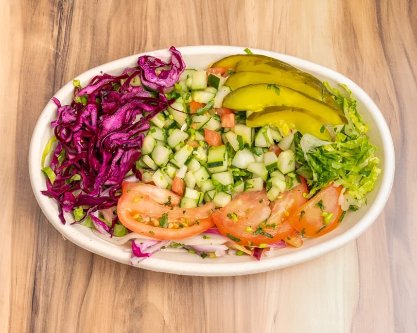 A vibrant salad with shredded red cabbage, diced cucumbers, sliced tomatoes, lettuce, and pickles.