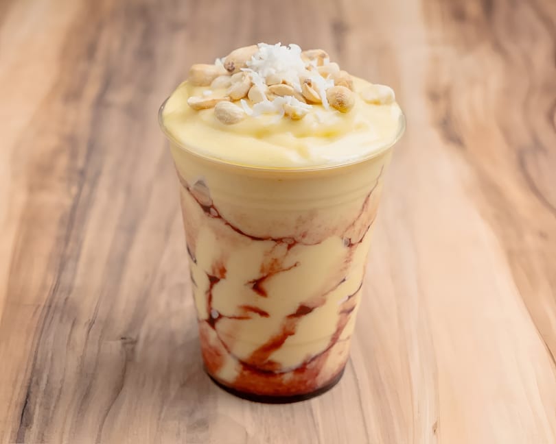 A plastic cup of yellow mango smoothie with red swirls, topped with peanuts and shredded coconut.
