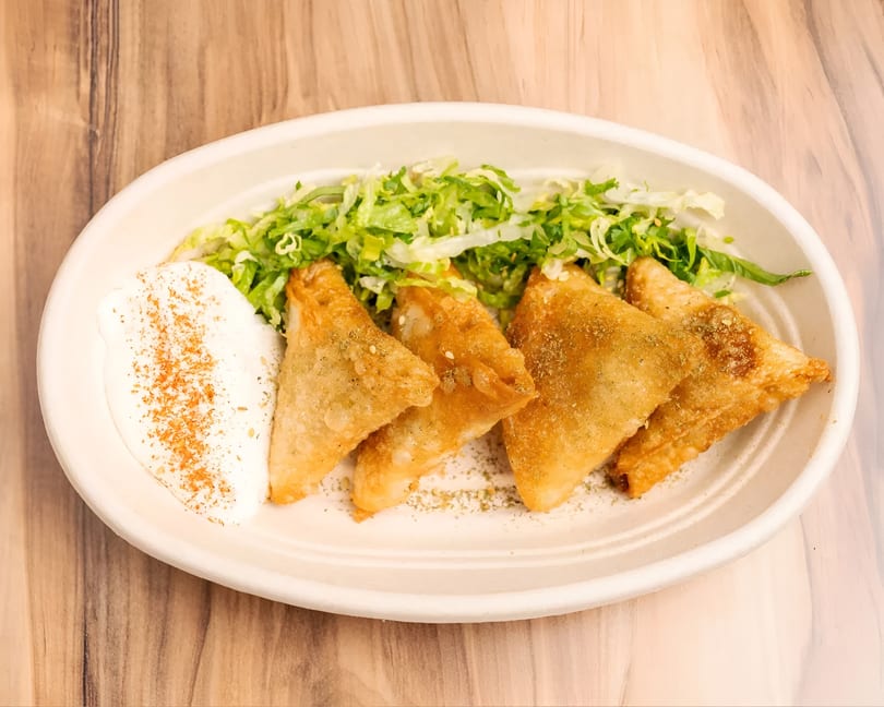 Four golden-brown samosas with a side of shredded lettuce and a dollop of white sauce.